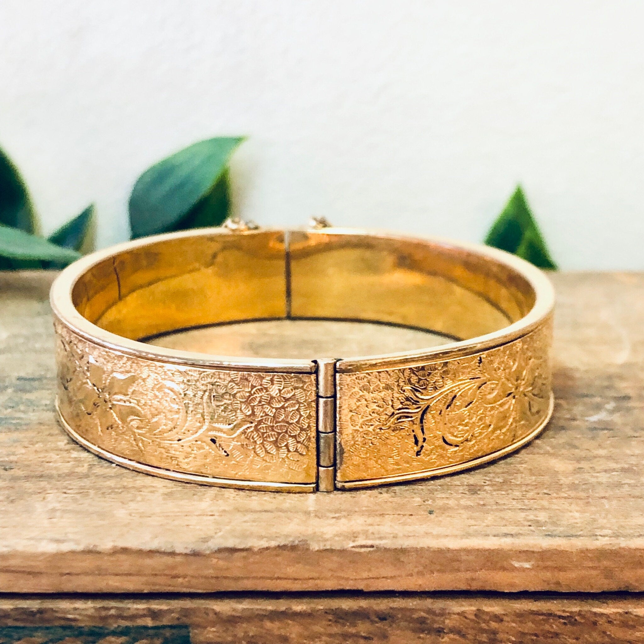 Vintage gold filled bangle bracelet with etched floral design on a wooden surface with leaves in the background.