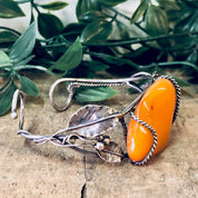 Vintage butterscotch amber bracelet with leaf design on a silver cuff, featuring orange amber stones and 925 silver, displayed on a natural background with foliage.