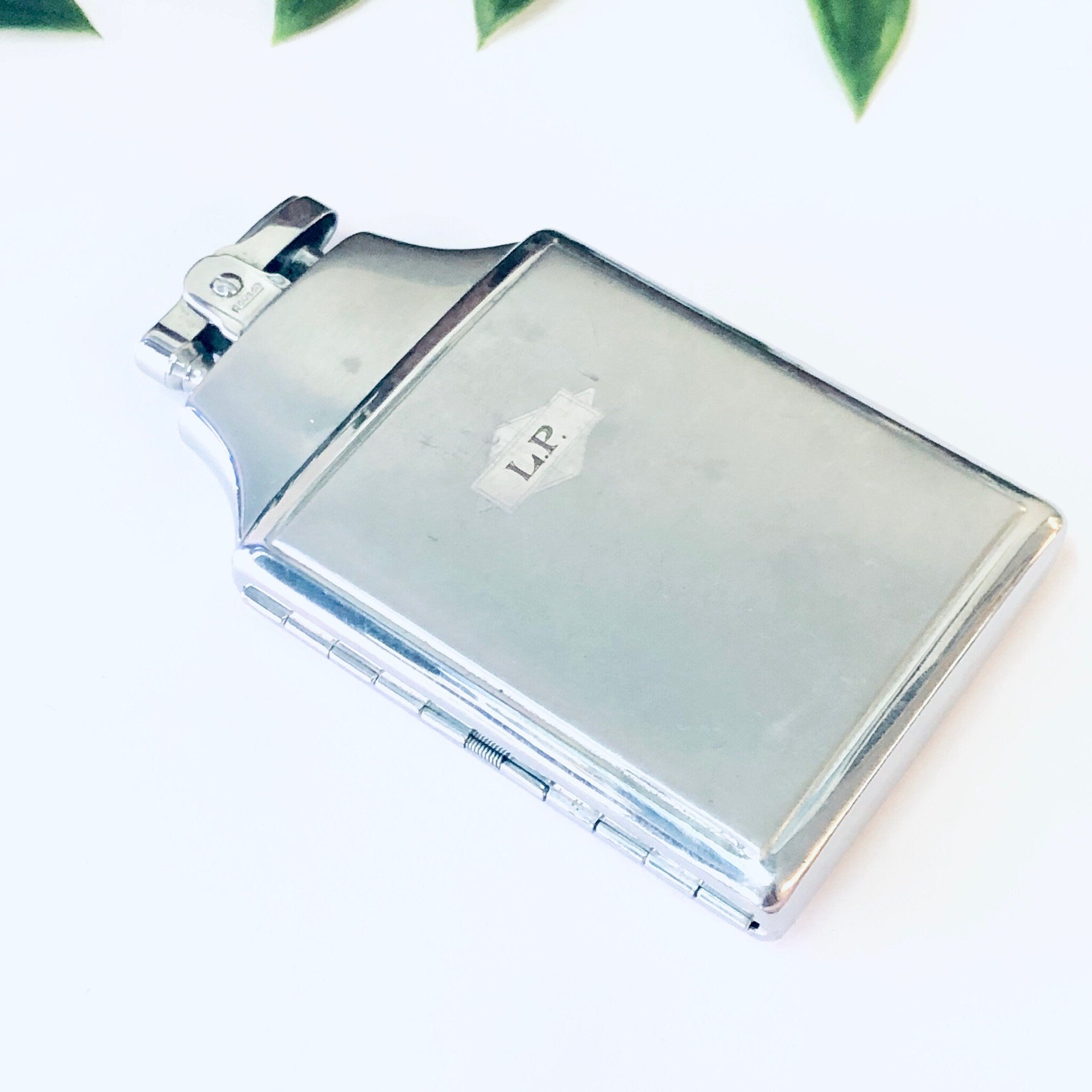 Vintage silver Ronson Mastercase cigarette holder and lighter with monogrammed initials, against a white background with green leaves at the top of the frame.