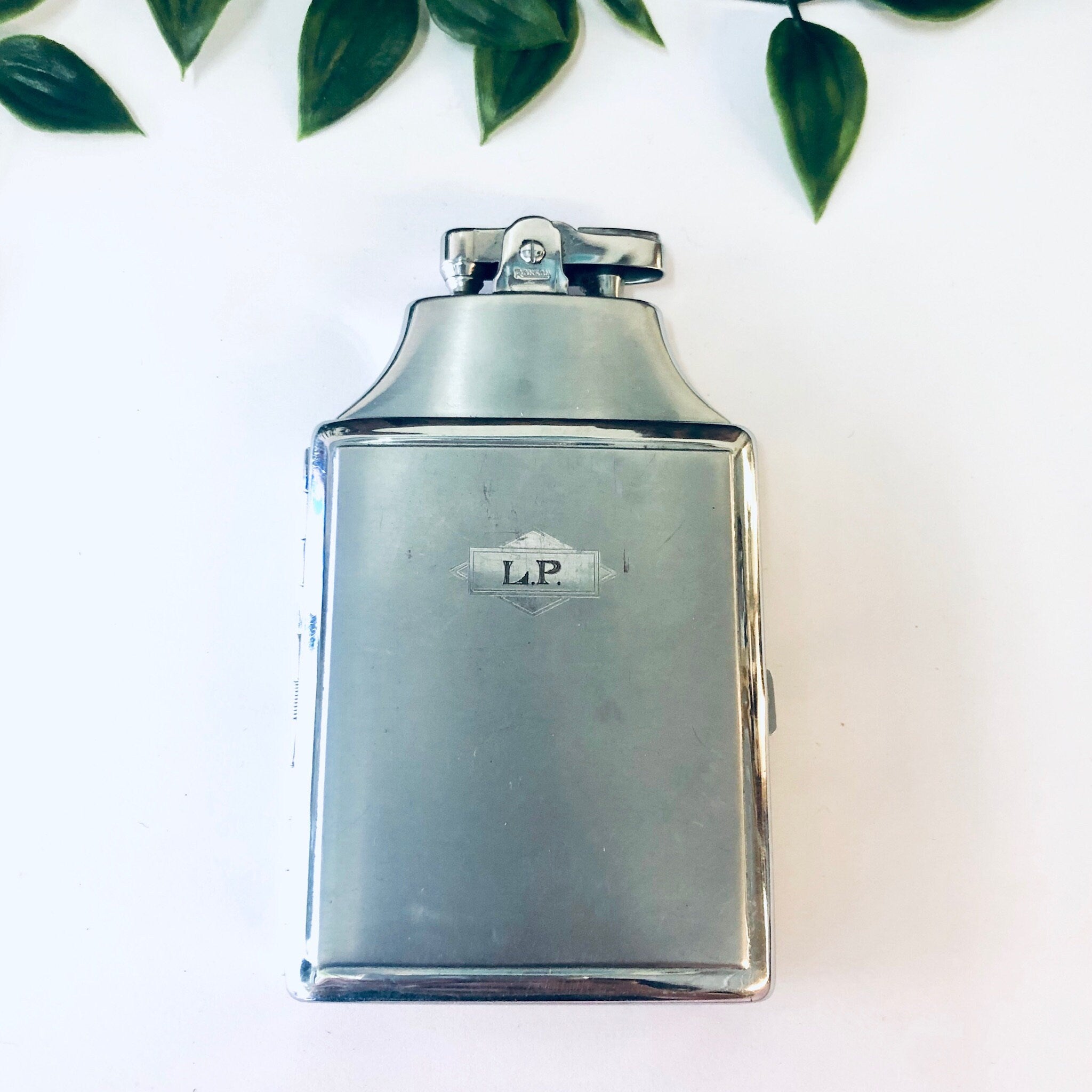 Vintage silver Ronson Mastercase cigarette holder and lighter with "LP" monogram, photographed against a white background with green leaves.
