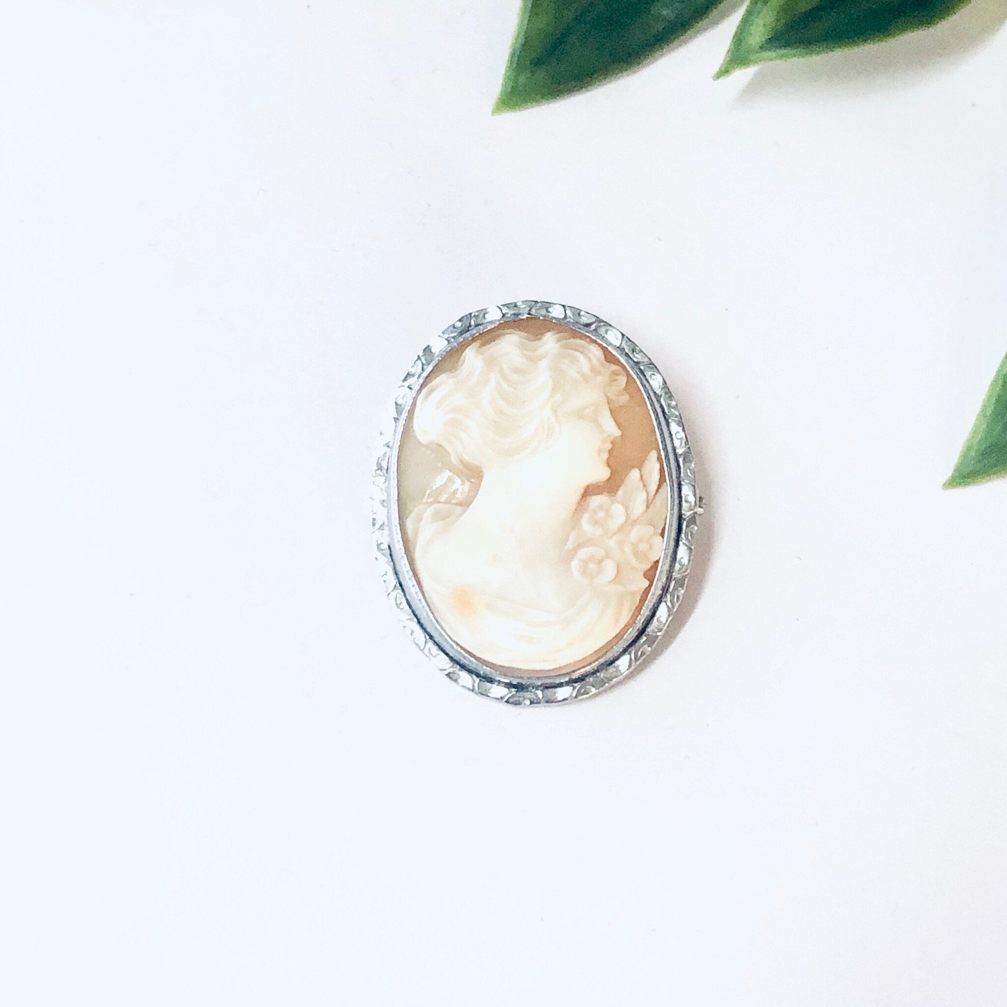 Vintage silver-toned cameo brooch pin featuring a carved ivory portrait set against green leaves on a white background.