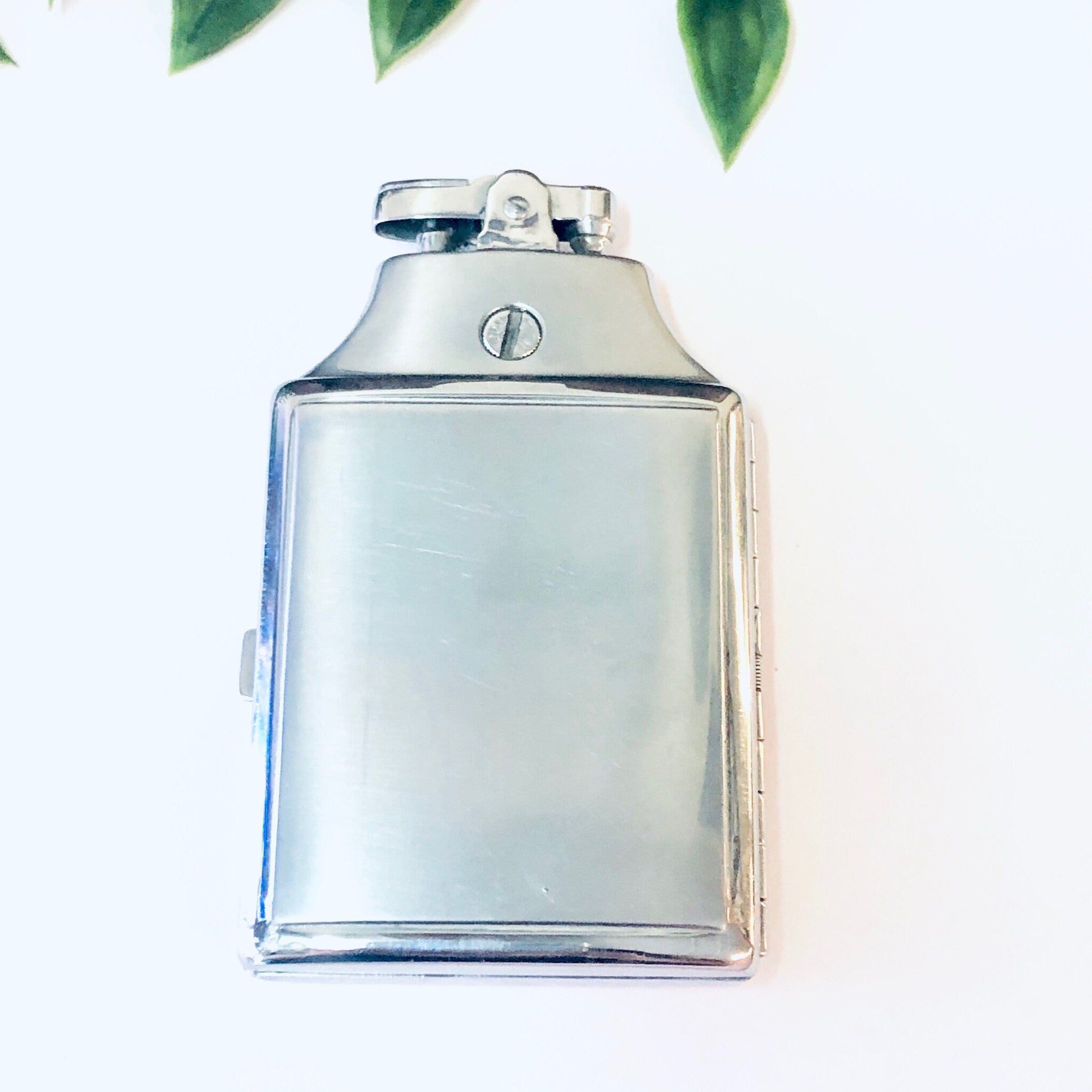 Vintage silver Ronson Mastercase cigarette lighter and holder, monogrammed, against a white background with green leaves.