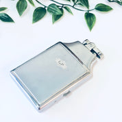 Vintage Ronson Mastercase silver cigarette holder and lighter with monogram initials, surrounded by green leaves on a white background