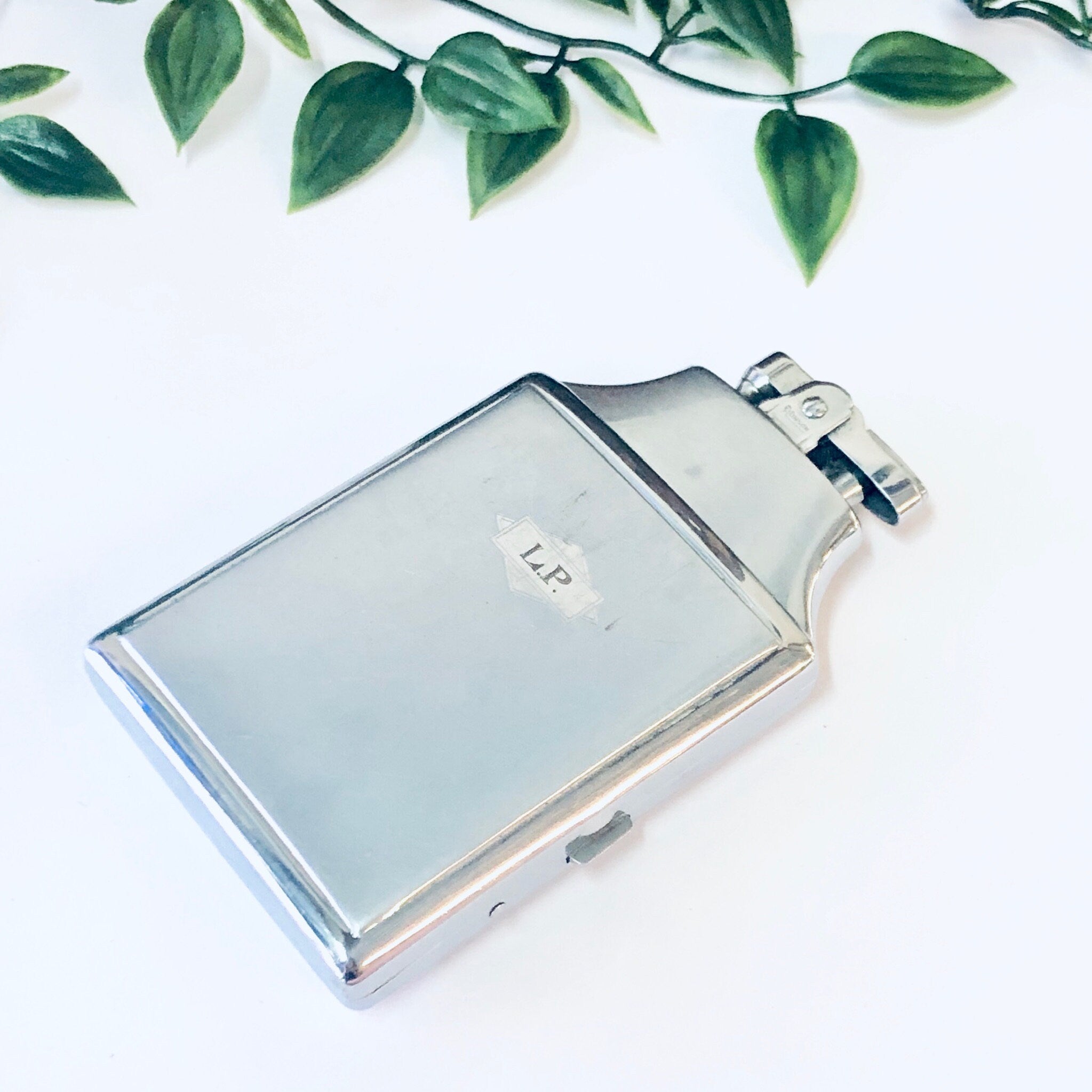 Vintage Ronson Mastercase silver cigarette holder and lighter with monogram initials, surrounded by green leaves on a white background