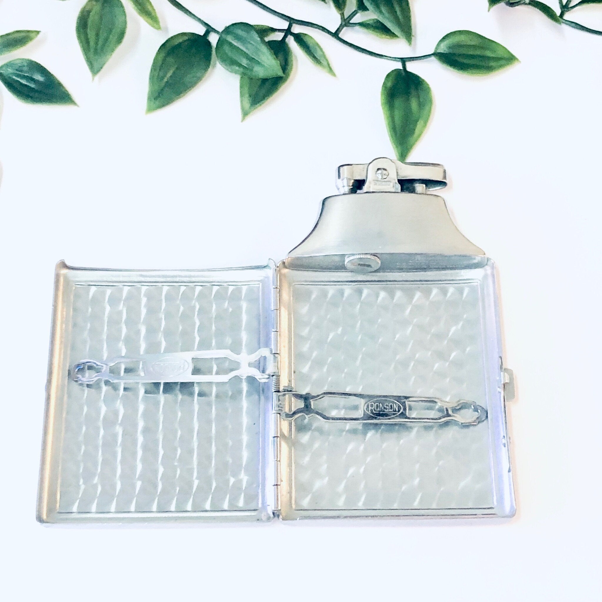 Vintage silver Ronson Mastercase cigarette holder and lighter with art deco design and monogram, displayed open with green leaves in the background.