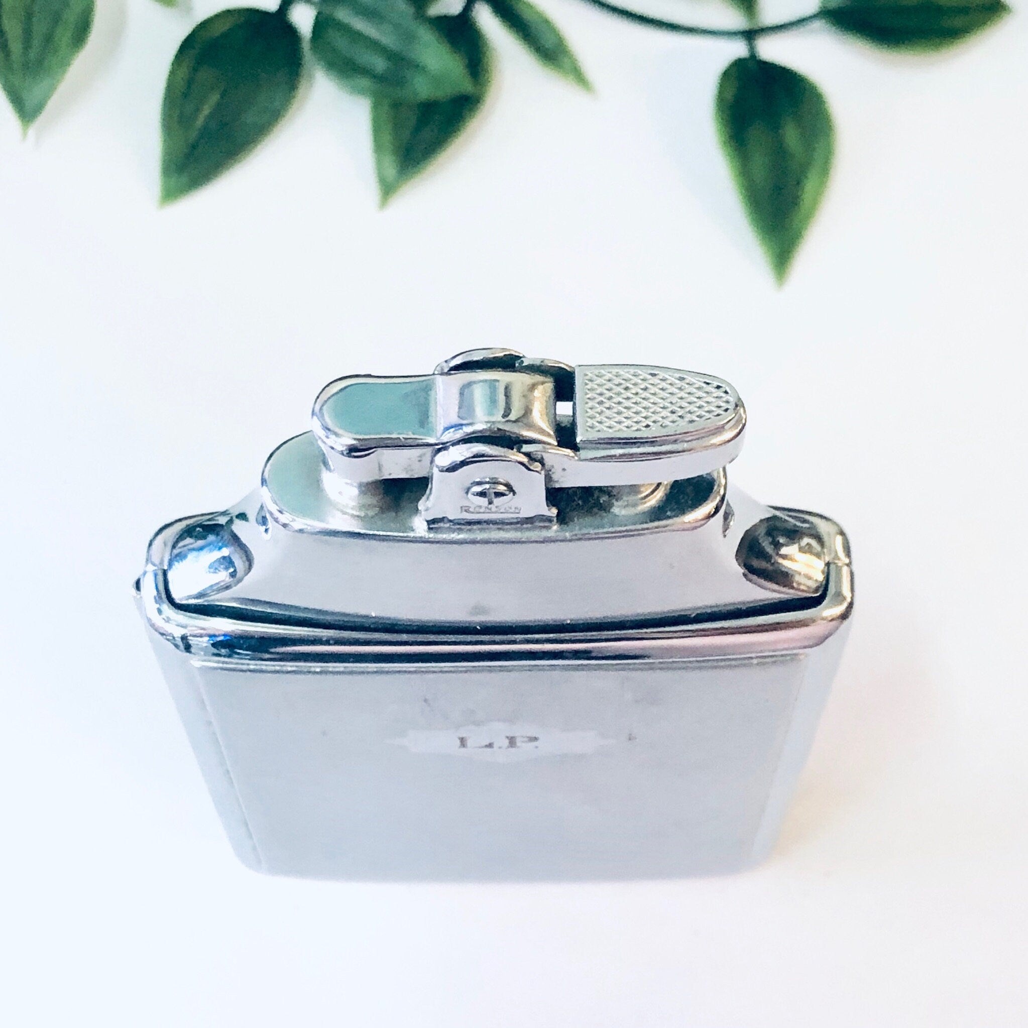 Vintage silver Ronson Mastercase cigarette lighter and holder with monogram, against a white background with green leaves.
