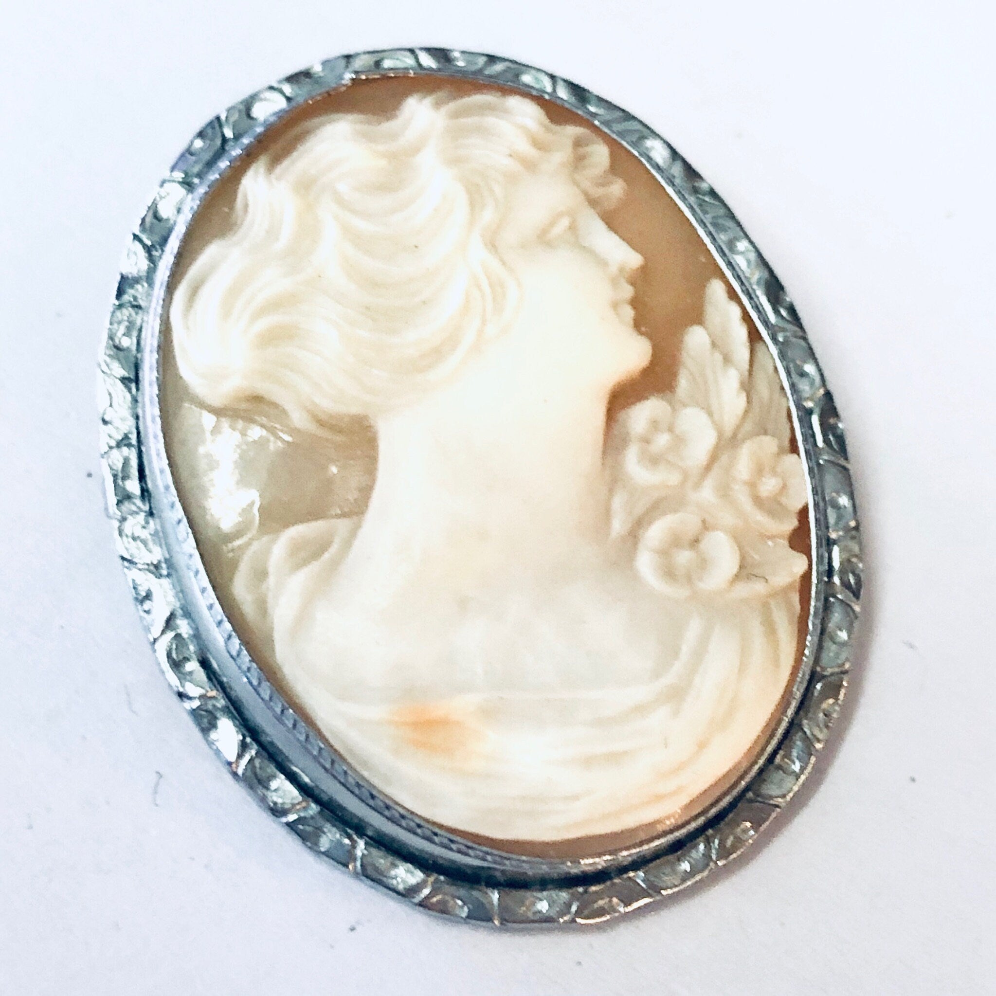 Vintage silver-toned cameo brooch pin featuring a detailed profile portrait carved in a pale oval stone, surrounded by an ornate floral border.