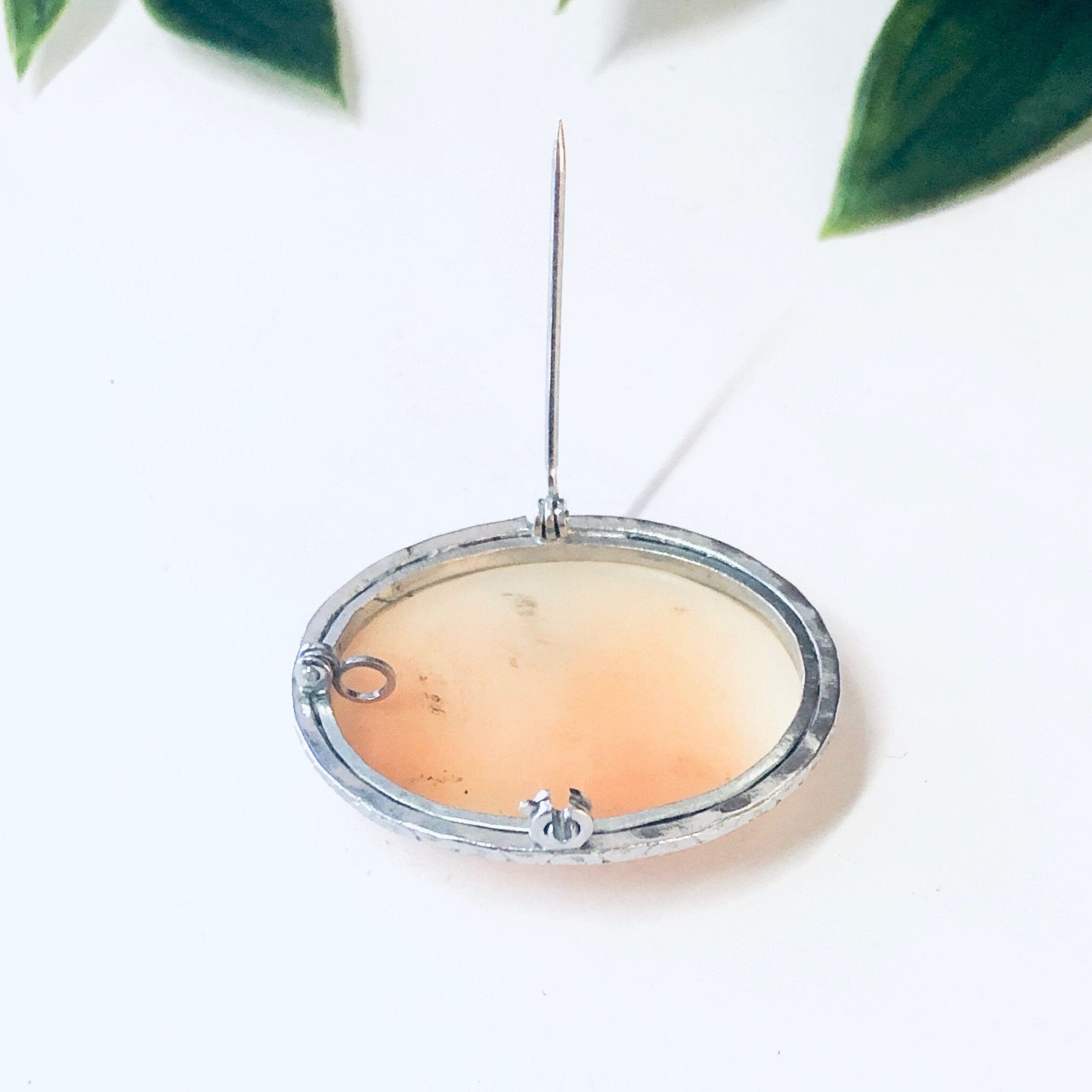 Vintage silver-toned cameo brooch pin pendant with oval peach stone set against green leaves on white background