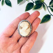 Vintage silver-toned cameo brooch pin held in hand with green leaves in the background