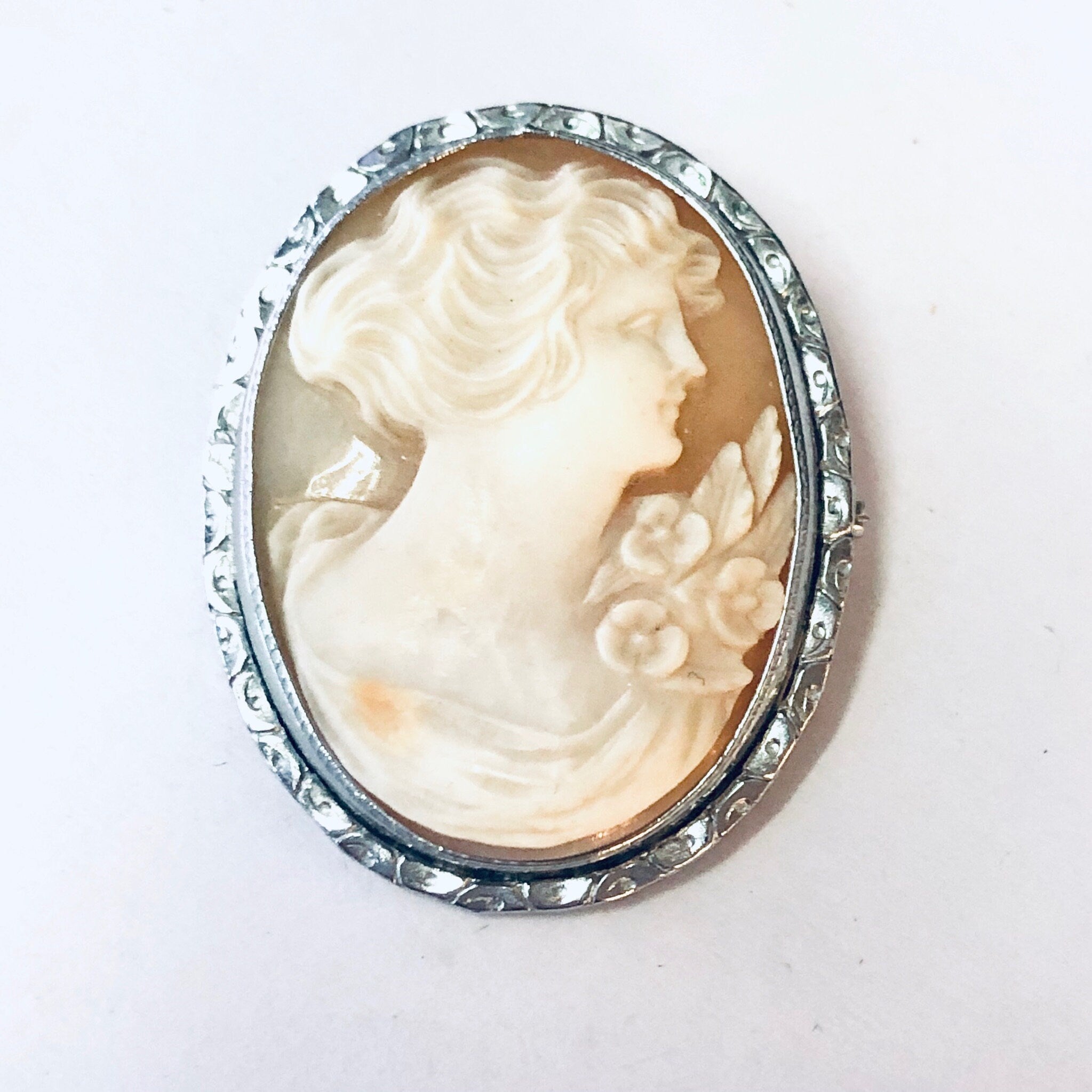Vintage silver-toned cameo brooch featuring a delicately carved female profile in a light-colored stone, surrounded by an ornate floral design border.