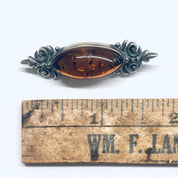 Vintage amber and silver brooch pin with ornate floral design, shown on an antique wooden ruler with "WM. F. LACY" text visible.