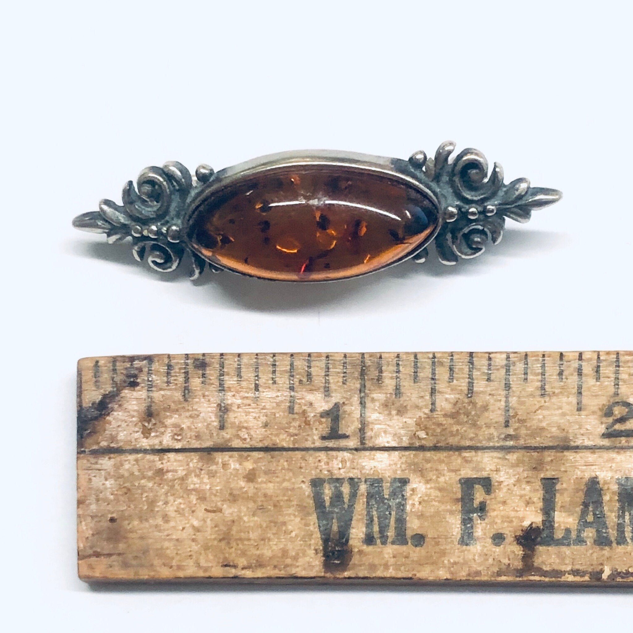 Vintage amber and silver brooch pin with ornate floral design, shown on an antique wooden ruler with "WM. F. LACY" text visible.