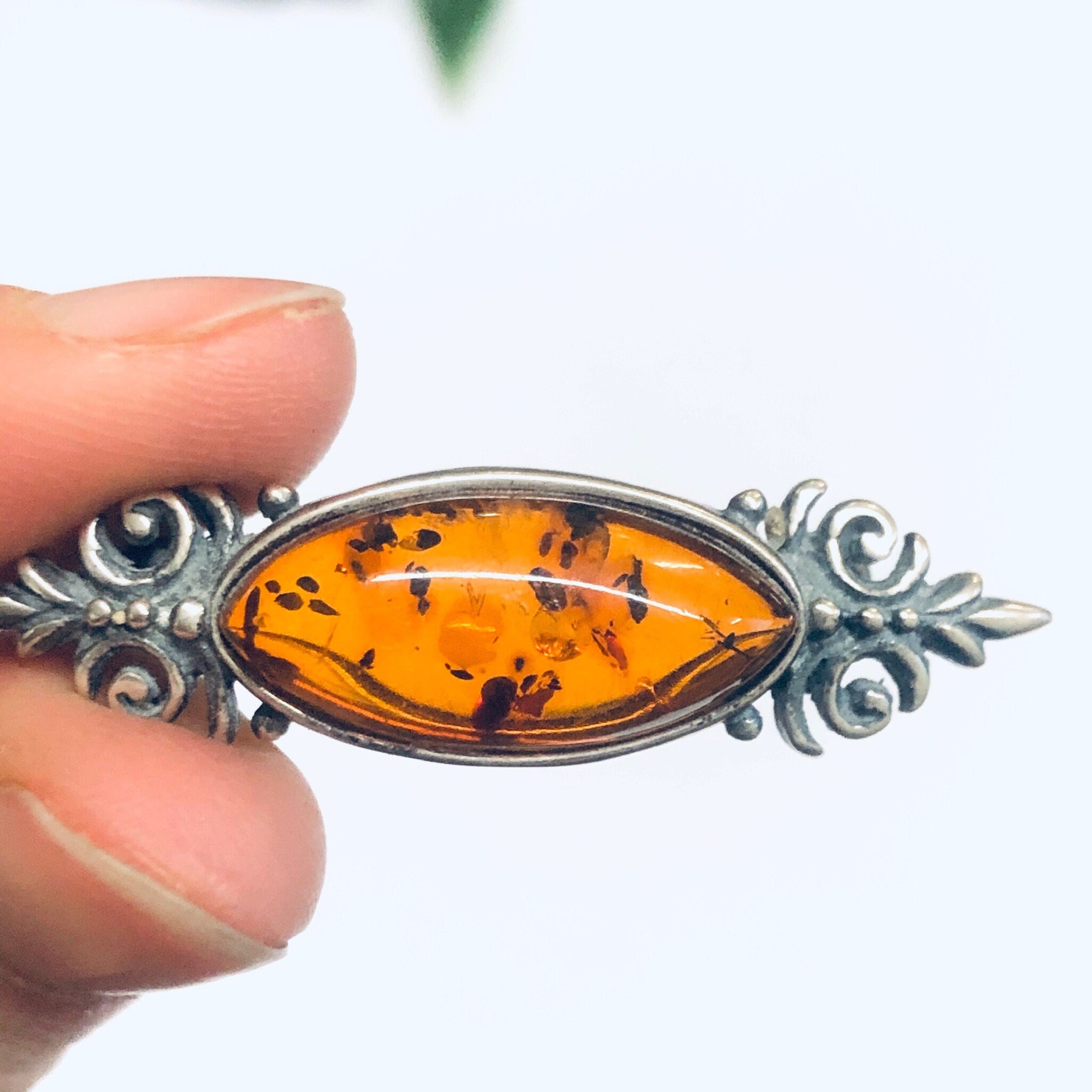 Vintage amber and silver brooch with intricate silver scrollwork holding an oval amber cabochon, held in fingers against a white background