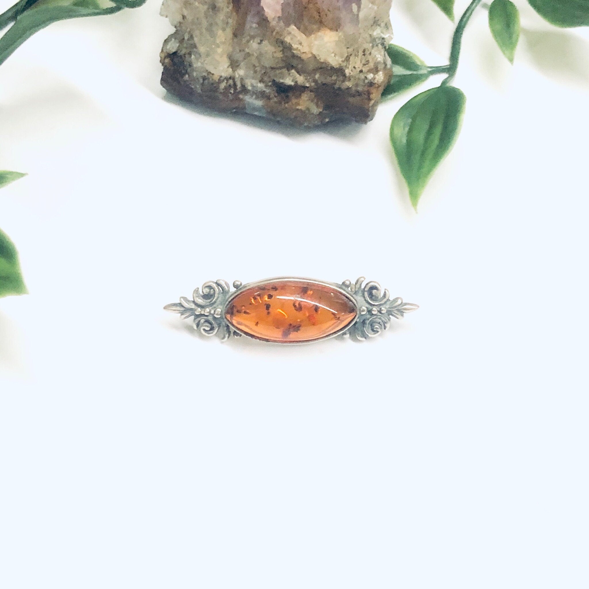 Vintage amber and silver brooch featuring an oval amber stone set in an ornate silver setting, displayed on a white background with greenery accents.