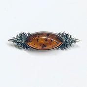 Vintage amber and silver brooch featuring an oval-shaped amber cabochon set in an ornate silver frame with floral and scroll designs, displayed on a white background.