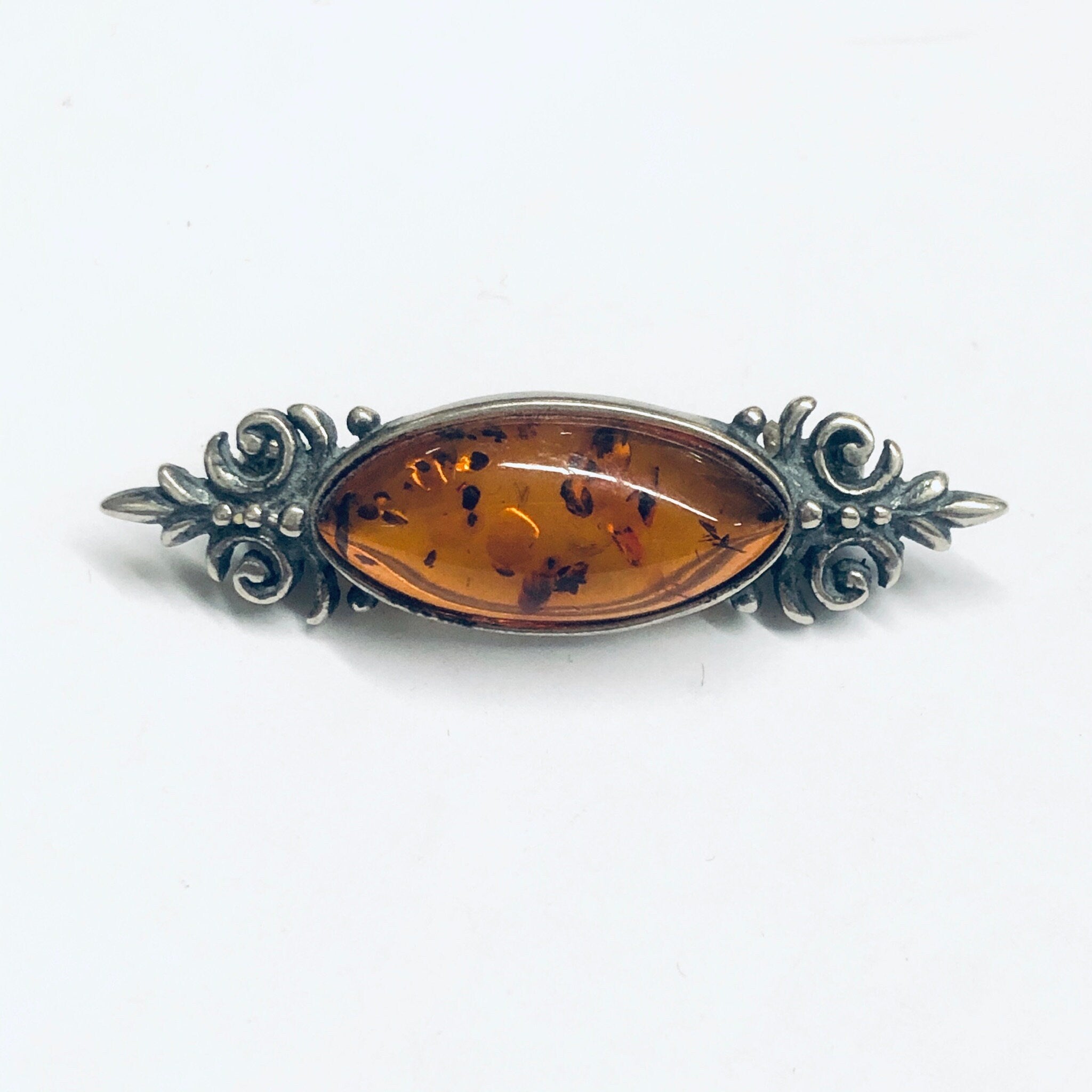 Vintage amber and silver brooch featuring an oval-shaped amber cabochon set in an ornate silver frame with floral and scroll designs, displayed on a white background.