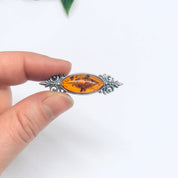 Vintage amber and silver brooch held between fingers, showcasing the small, unique amber stone set in an ornate silver setting against a plain white background.