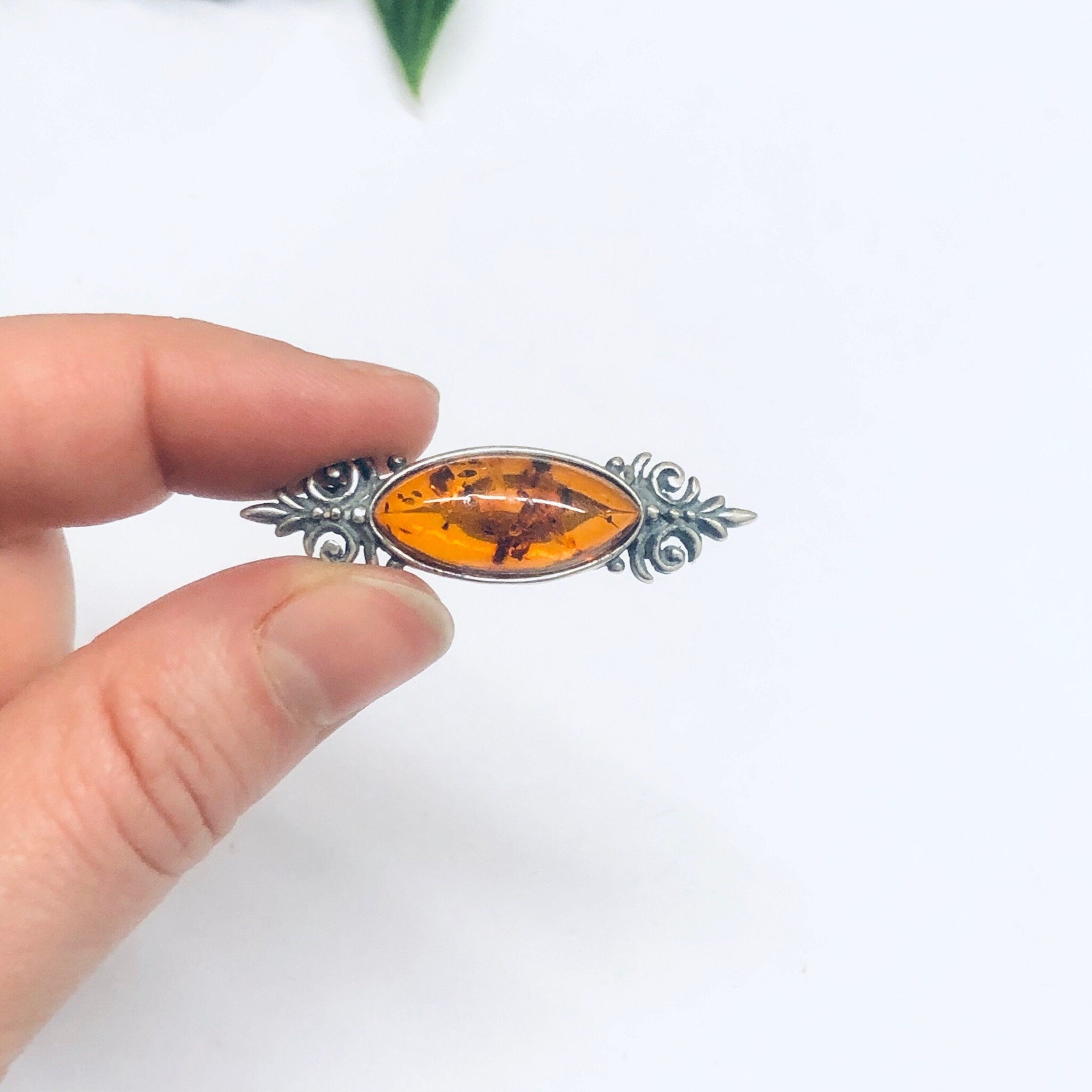 Vintage amber and silver brooch held between fingers, showcasing the small, unique amber stone set in an ornate silver setting against a plain white background.