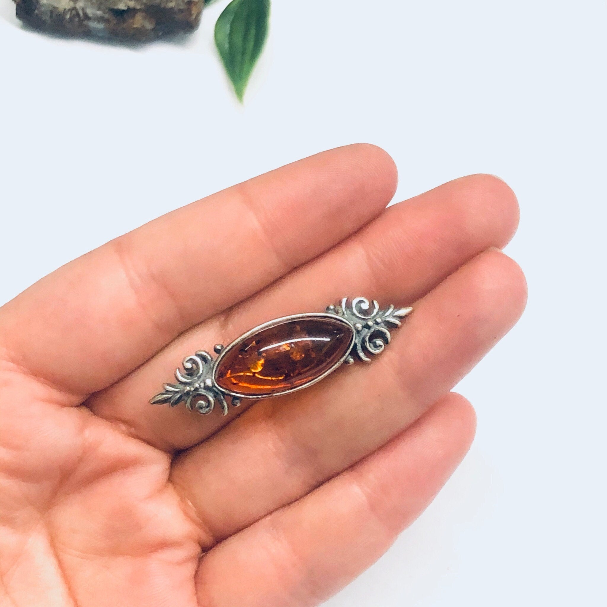 Vintage silver brooch with amber stone held in an ornate floral setting, shown in a person's hand to demonstrate the small size of the unique amber jewelry piece.