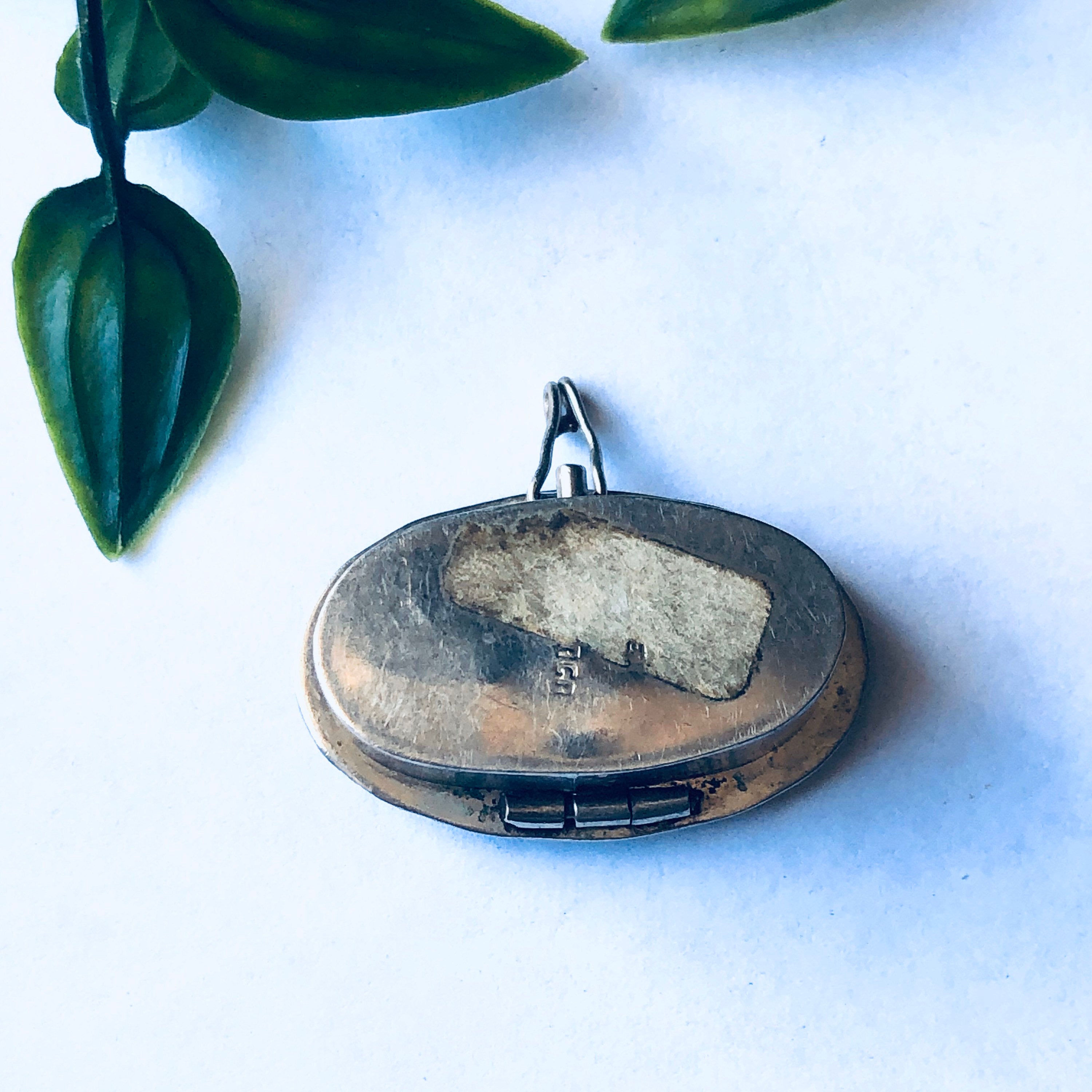 Small silver pill box with abalone shell inlay, vintage accessory for holding pills or small items, adorned with iridescent blue and green shell pieces, set against a white background with green leaves.