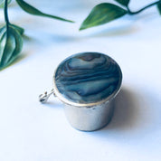 Vintage silver pill box with blue and green abalone shell inlay, small round shape, displayed with greenery on a white background.
