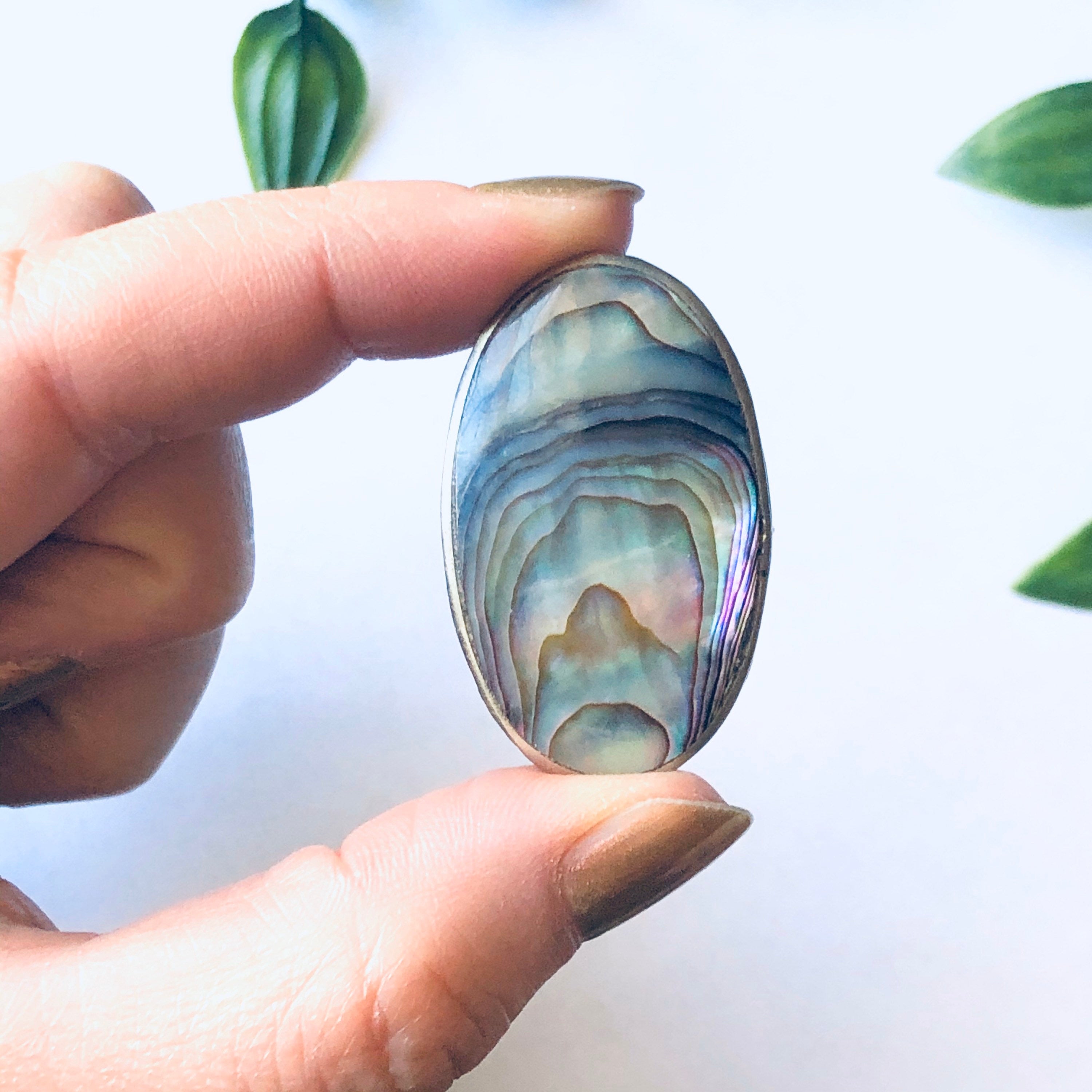 Small vintage silver pill box with blue and green abalone shell inlay held between fingers with leaves in background
