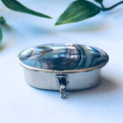 Small vintage silver pill box with blue and green abalone shell inlay, displaying an oval shape with legs, photographed with green leaves in the background.