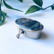 Small vintage silver pill box with abalone shell inlay, featuring iridescent blue and green colors, against a white background with green leaves.