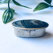 Small vintage silver pill box with blue and green abalone shell inlay, set against green leaves on a white background.