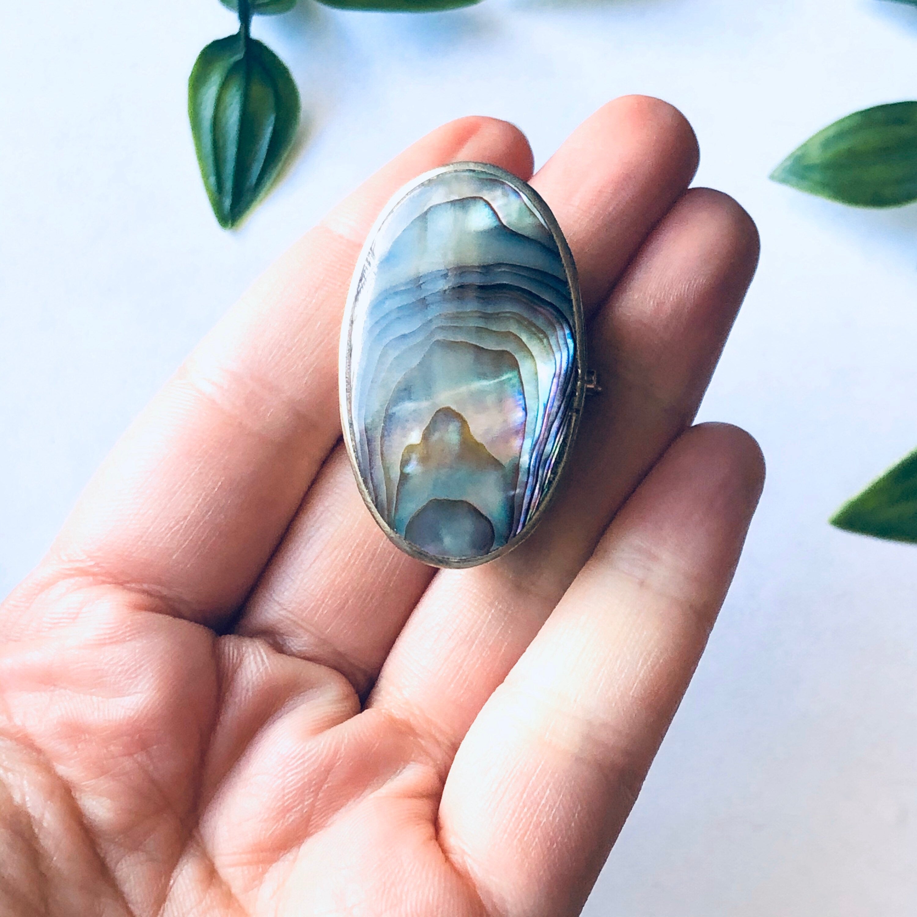 Small vintage silver pill box held in hand, decorated with iridescent blue and green abalone shell inlay, against white background with green leaves.