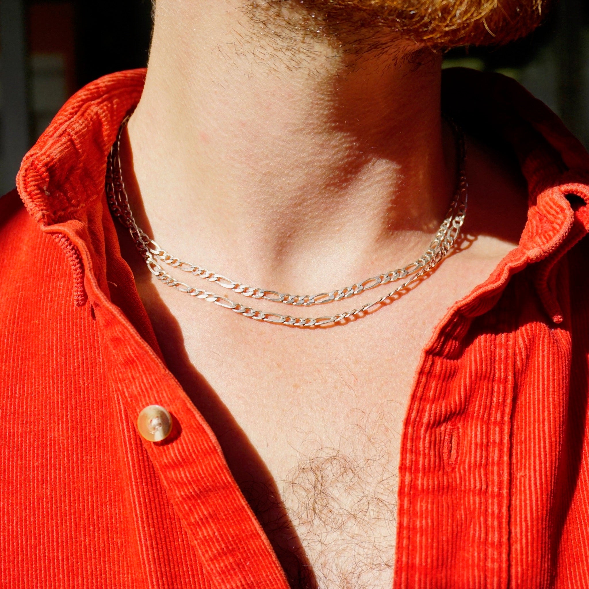 Closeup of man's neck wearing a vintage sterling silver extra long 34 inch figaro chain necklace made in Italy, layered over red corduroy shirt, against blurred background.