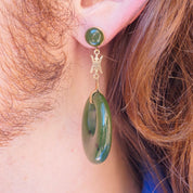 14K gold dangle earrings with natural green jade hoops and Chinese symbol charms, worn on the ear of a person with long brown hair.