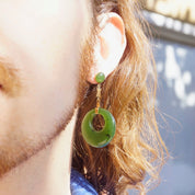 Close-up of a woman's ear with long blonde hair wearing 14K gold dangle earrings featuring green jade hoops suspended from a Chinese symbol charm.