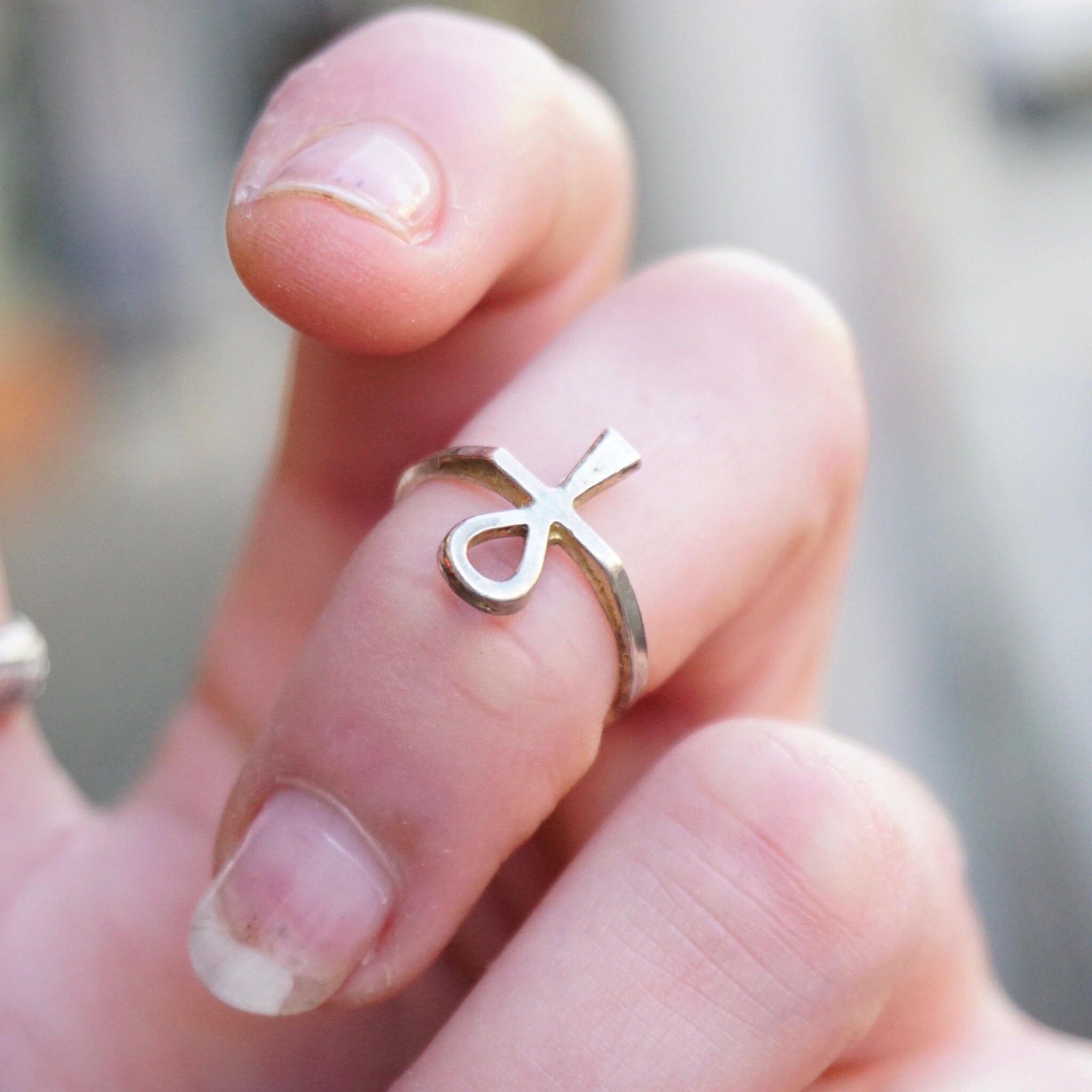 Close-up view of a hand wearing a thin, silver ankh ring on the ring finger. The ankh, an ancient Egyptian hieroglyphic symbol, is the focal point of the delicate sterling silver ring.