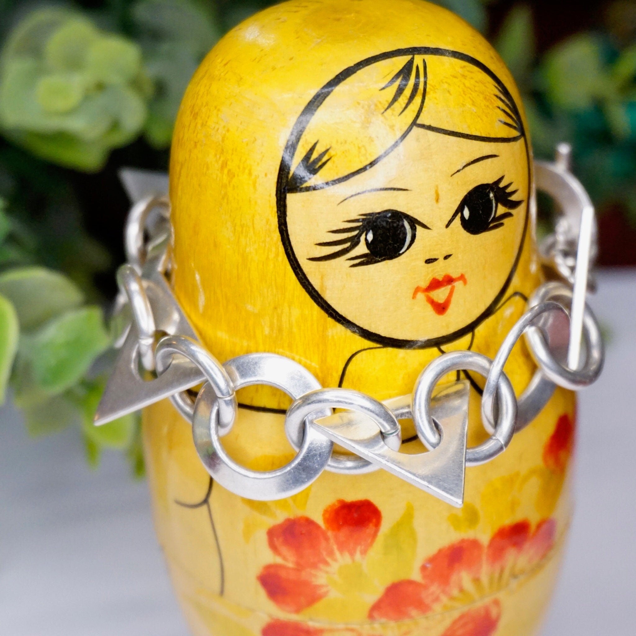 Painted wooden Russian nesting doll with blonde hair, rosy cheeks and large cartoon eyes, wearing a floral dress, chained together with metal links on a blurred background of succulent plants.