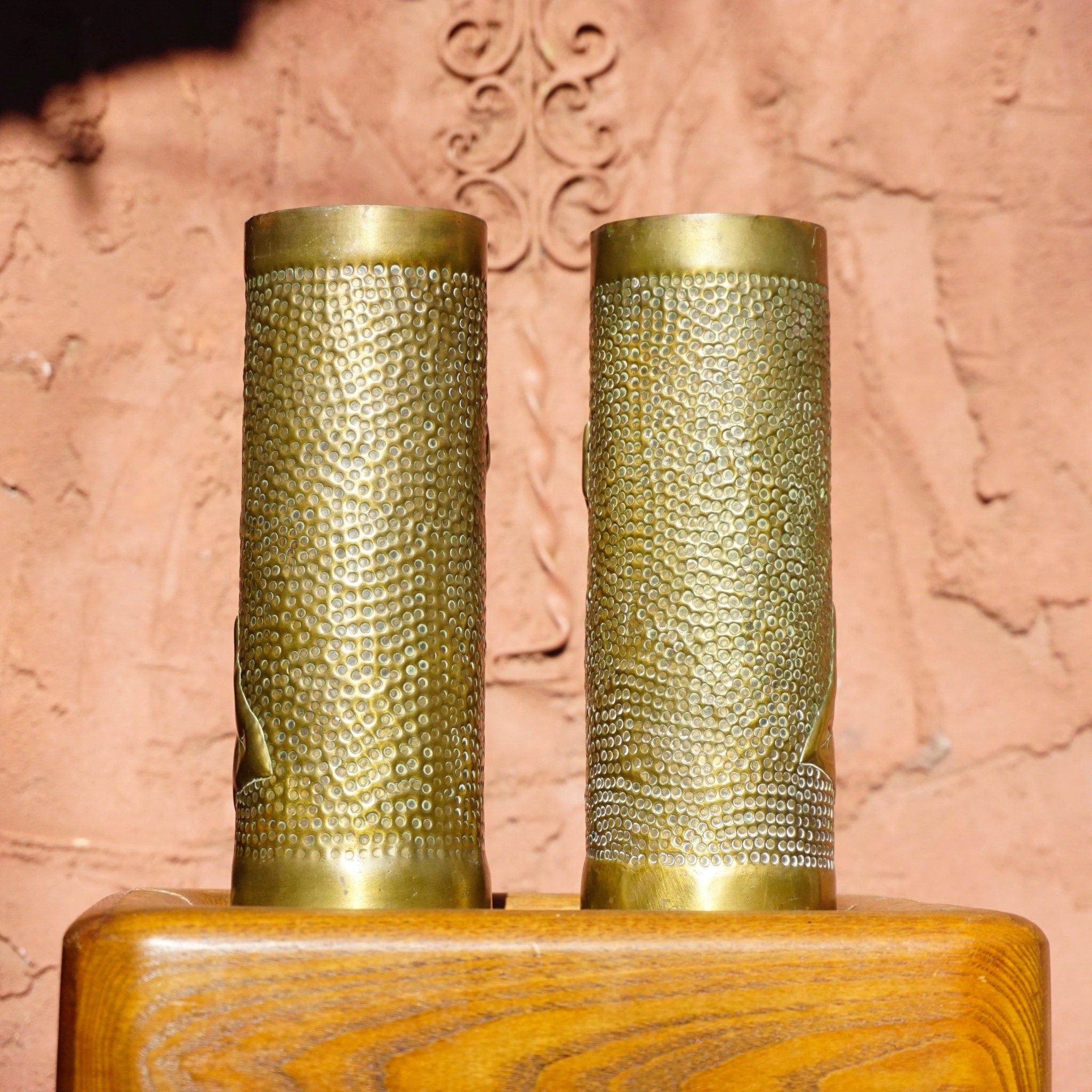 Antique French WWI trench art brass shell casing brush holders from 1917 with etched leaf design, standing on wooden surface against ornate pink wall