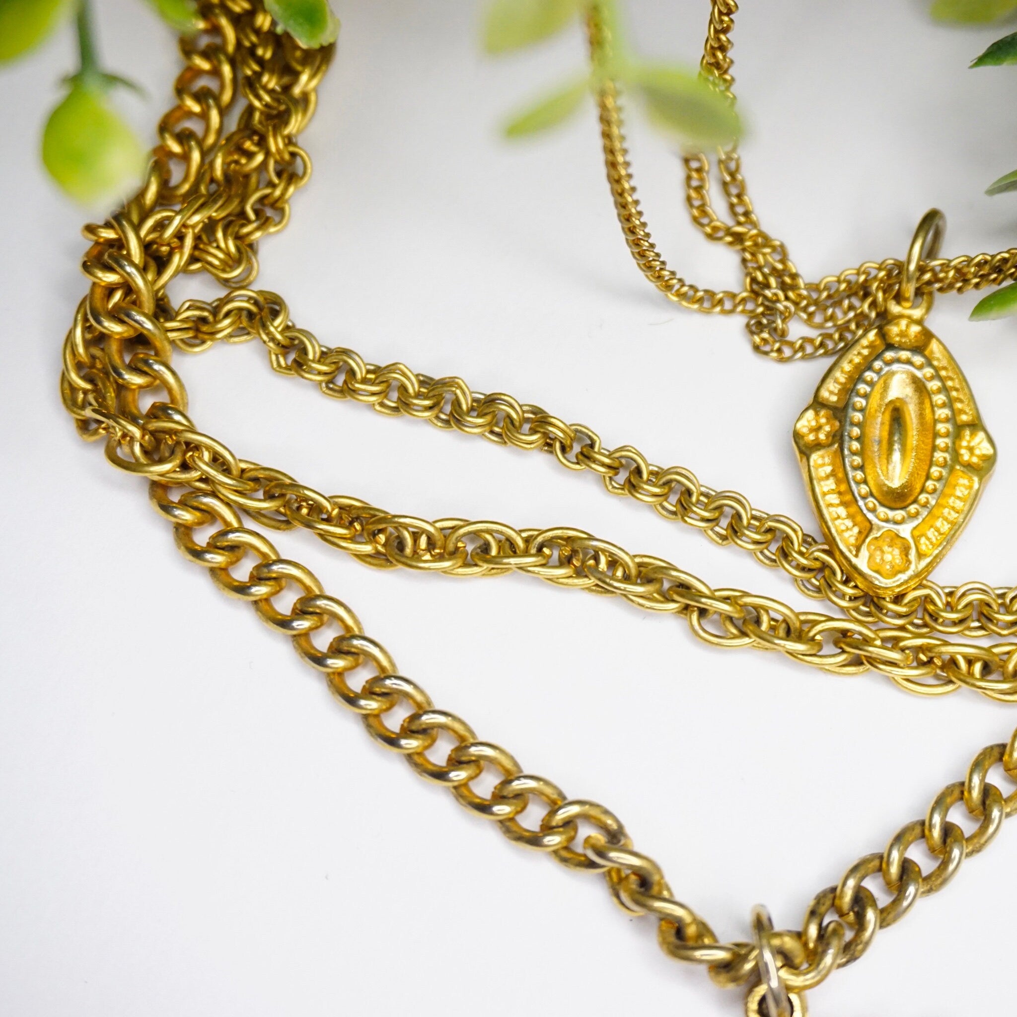 Vintage gold tone layered chain necklace featuring multiple strands and ornate gold pendants, set against a white background with green foliage accents.
