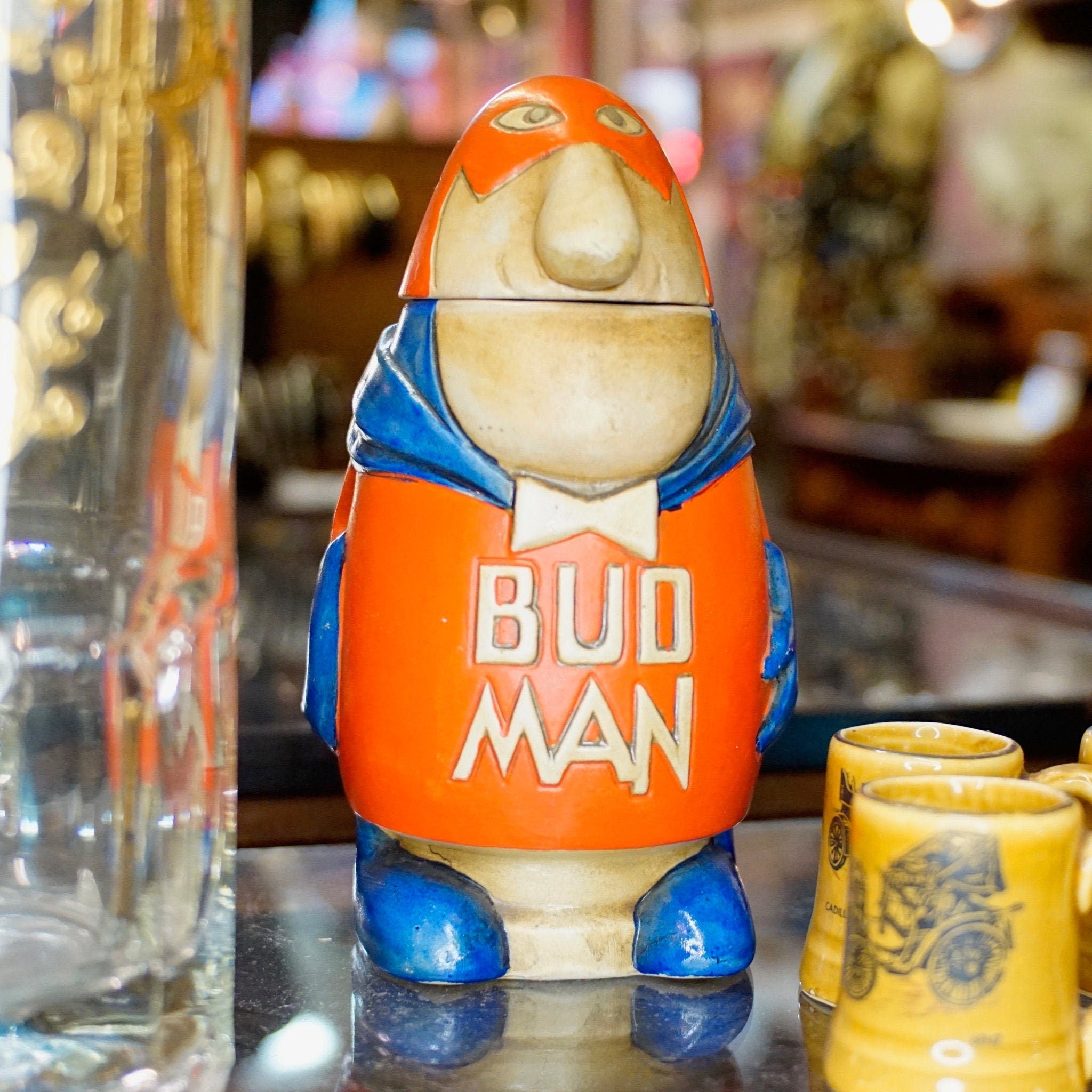 Vintage 1975 Bud Man beer stein figurine with orange jumpsuit and blue boots, standing on bar counter next to shot glasses.