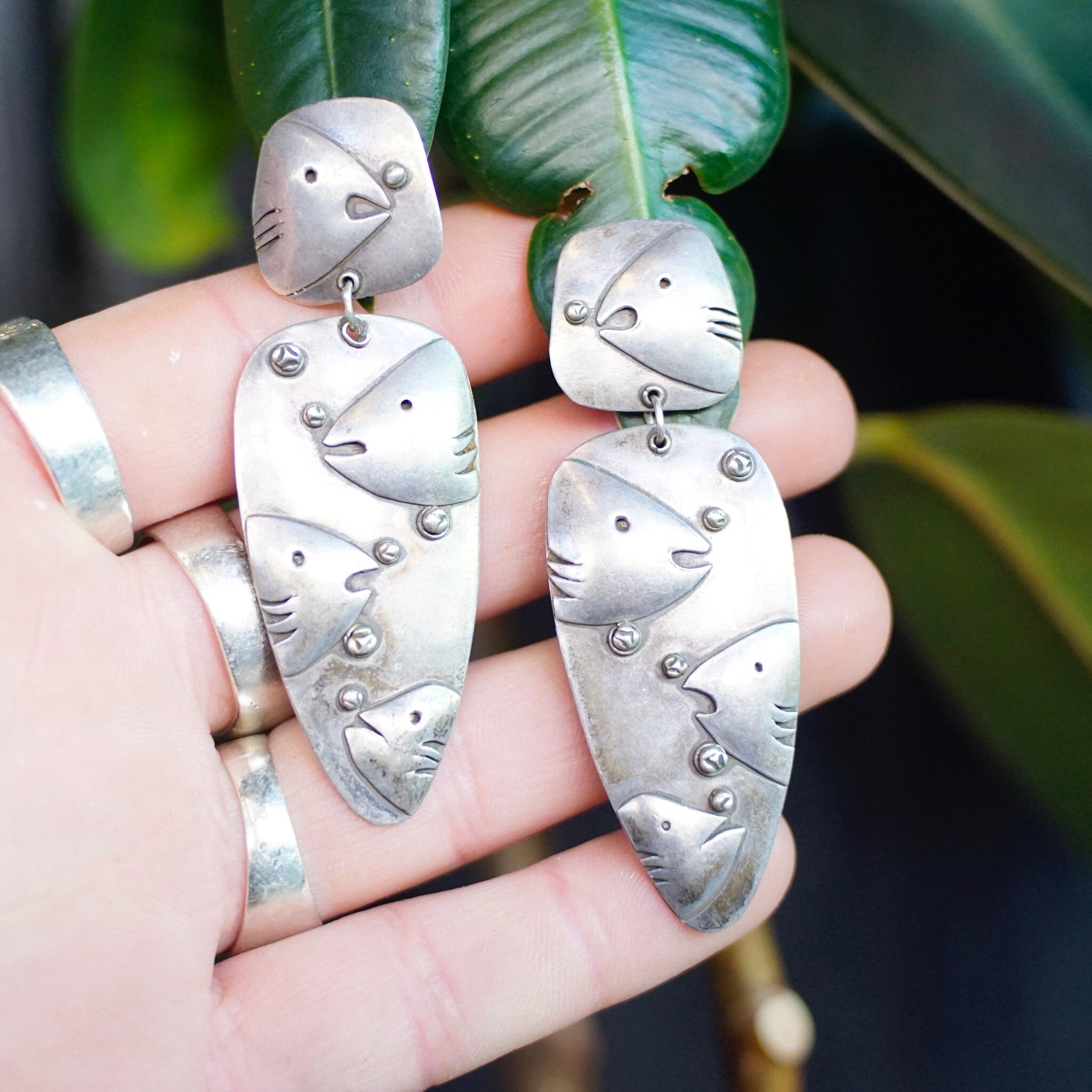 Vintage sterling silver embossed fish and bubble design dangle earrings held in palm of hand with leaves in background