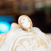 Antique 14K gold cameo ring with carved shell silhouette, ornate gold setting and accent diamond, shown resting on a textured white surface.