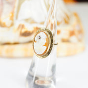 Closeup of an antique 14K gold cameo ring with an ornate setting and a small accent diamond, the carved shell cameo depicting a woman's side profile, shown against a blurred background.
