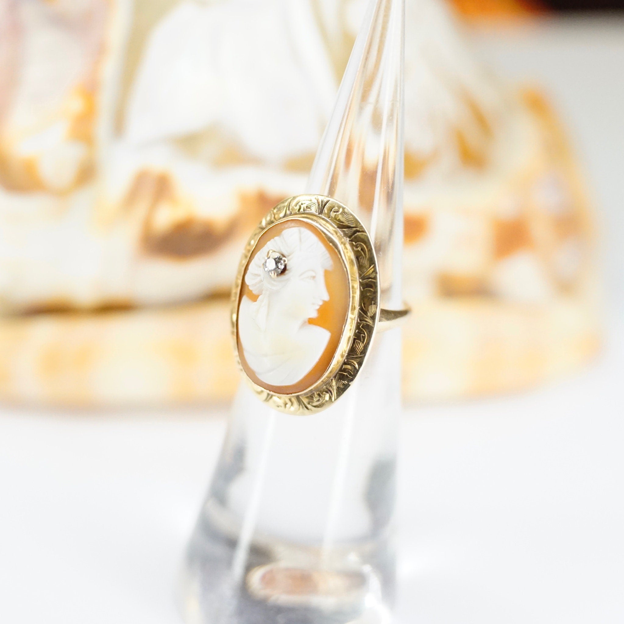Closeup of an antique 14K gold cameo ring with an ornate setting and a small accent diamond, the carved shell cameo depicting a woman's side profile, shown against a blurred background.
