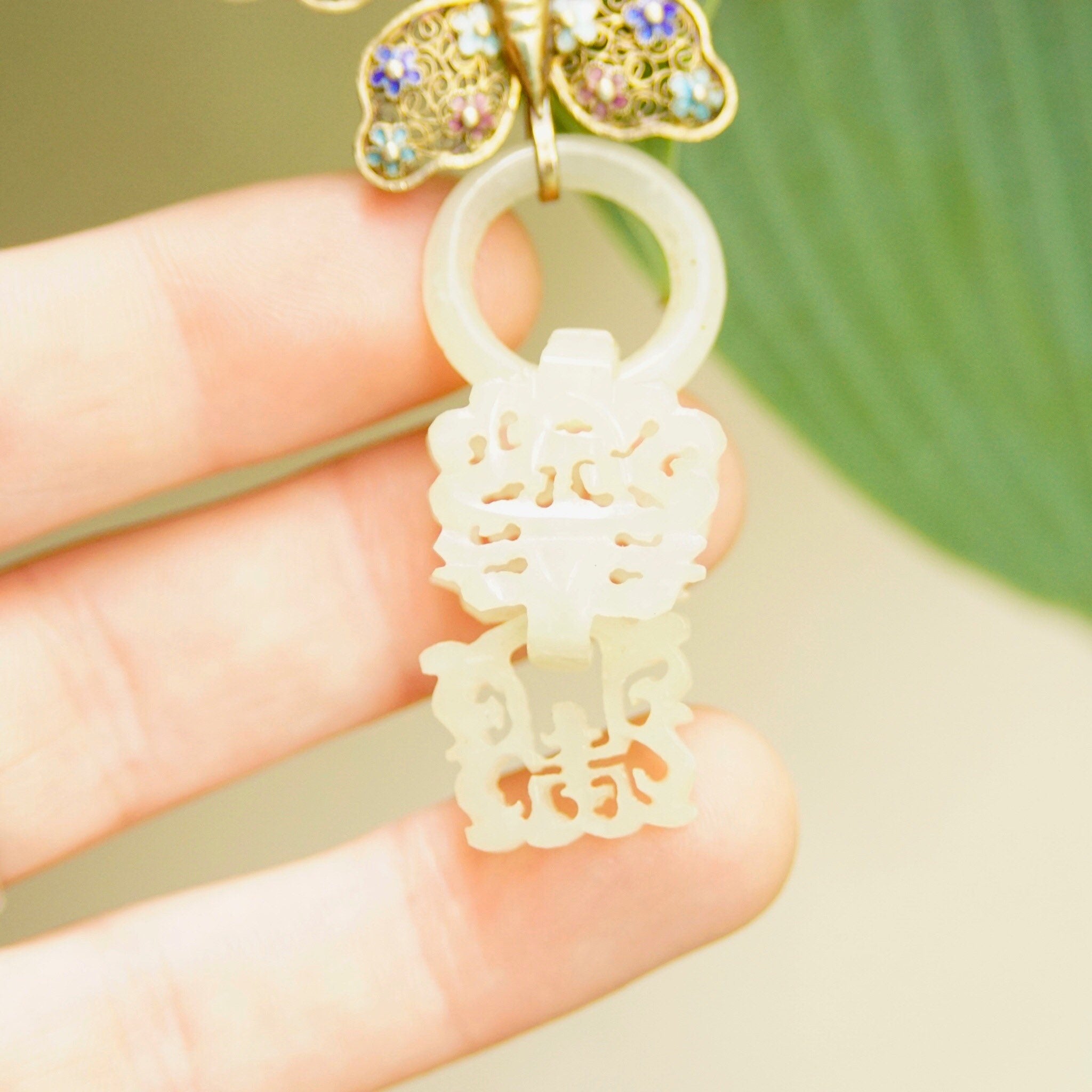 Antique Chinese silver filigree devil's work nephrite jade butterfly brooch pin with gold wash and hand-carved jade wedding basket charm, held in a person's fingers to show intricate details and craftsmanship.