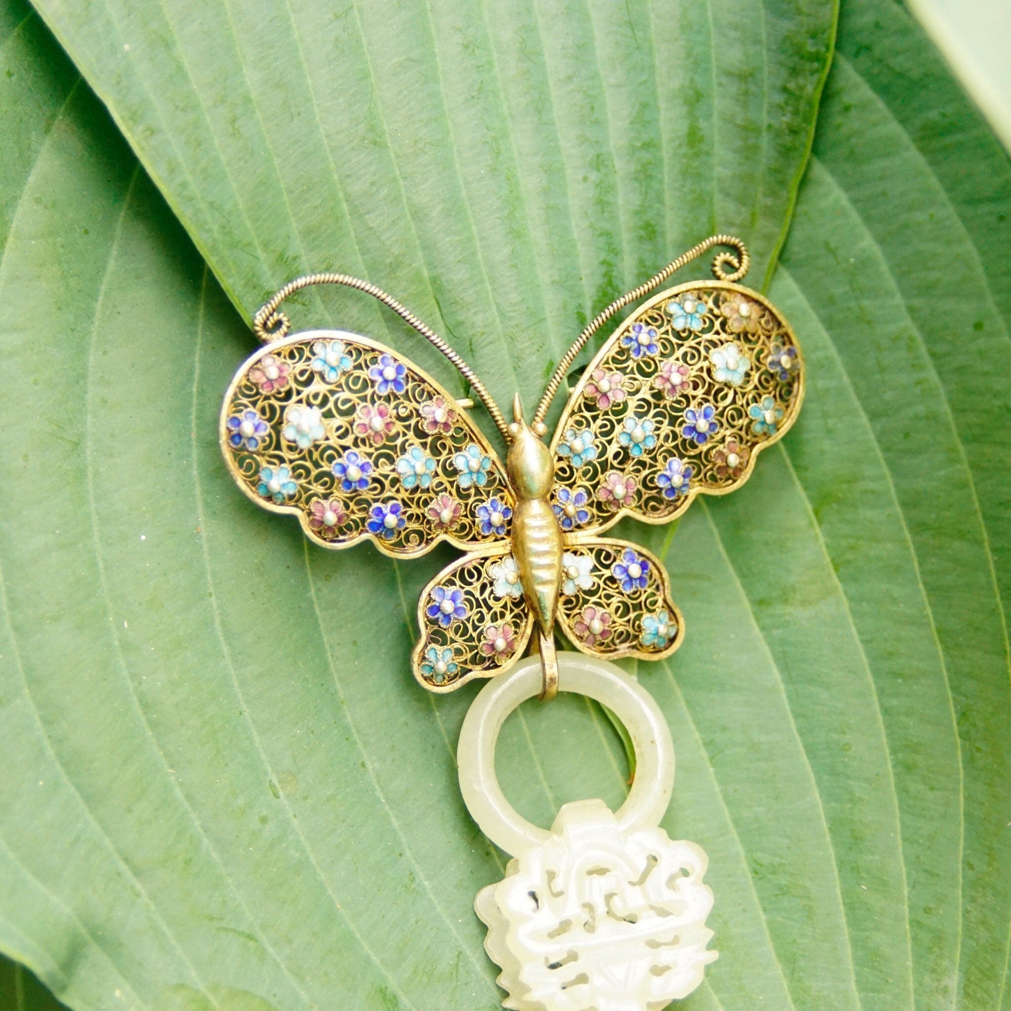 Alt text: Antique Chinese silver filigree devil's work nephrite jade butterfly brooch with gold wash, hand-carved jade wedding basket charm, set against green leaf background.
