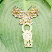Antique Chinese silver filigree butterfly brooch with gold wash and intricate devil's work design, featuring a hand-carved nephrite jade wedding basket charm dangling from the center, set against a green leaf background.