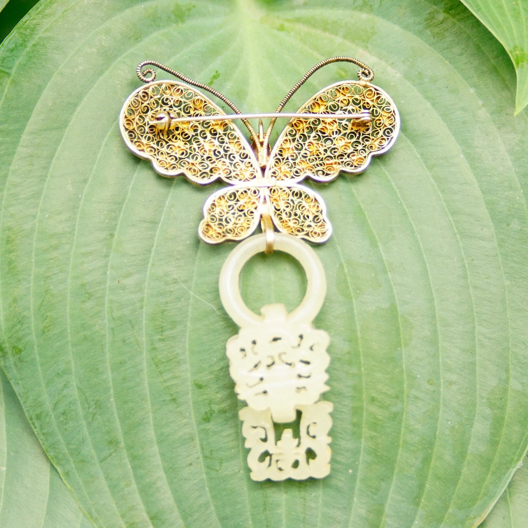Antique Chinese silver filigree butterfly brooch with gold wash and intricate devil's work design, featuring a hand-carved nephrite jade wedding basket charm dangling from the center, set against a green leaf background.