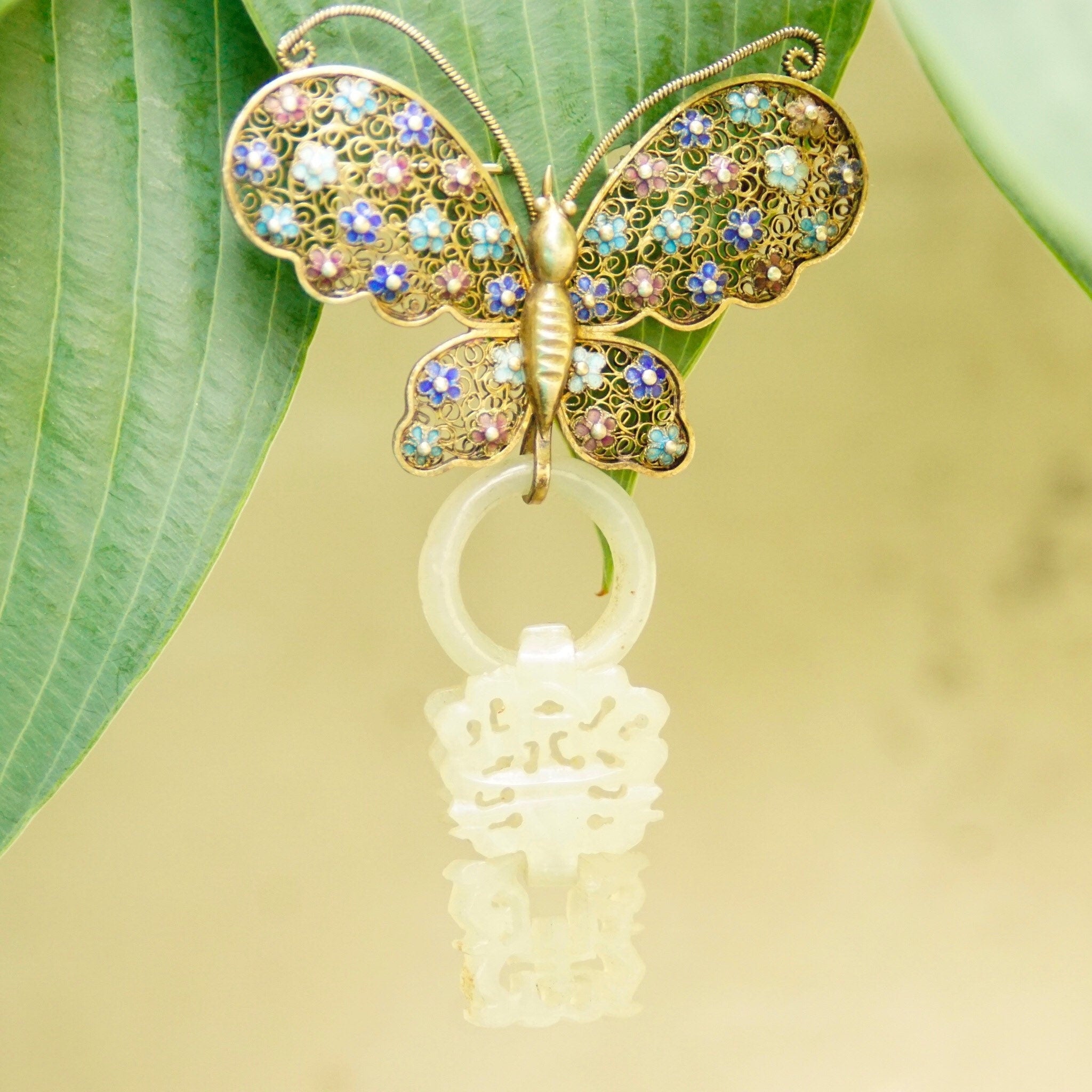 Antique Chinese silver filigree butterfly brooch with gold wash, enamel accents and a hand-carved nephrite jade wedding basket charm dangling from the bottom. The intricate silver openwork is accented with small blue, pink and green enamel flowers. The jade charm is a delicate, pale green color carved in the shape of a traditional Chinese wedding basket.