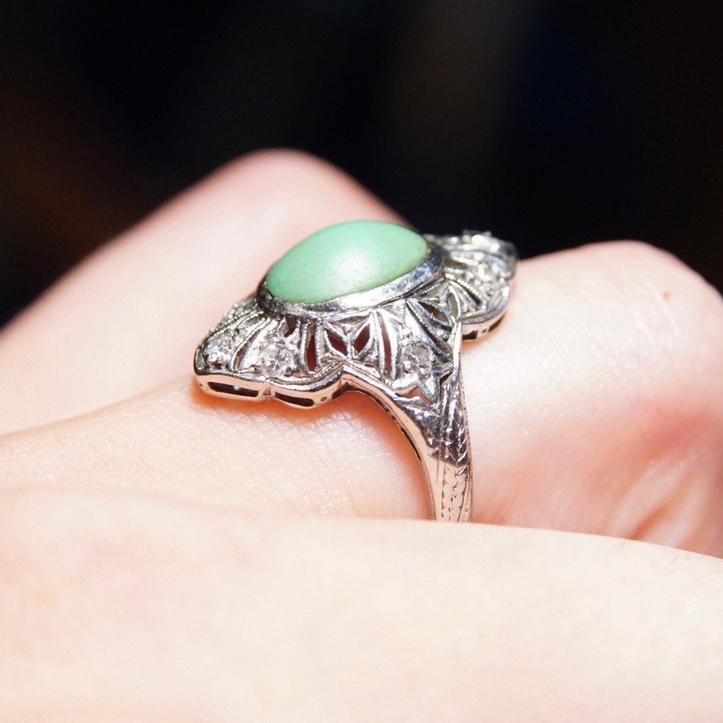 Vintage 18K white gold Art Deco filigree ring with matte green turquoise stone and diamond encrusted shield design, size 7 3/4 US, shown on a person's finger against a black background.