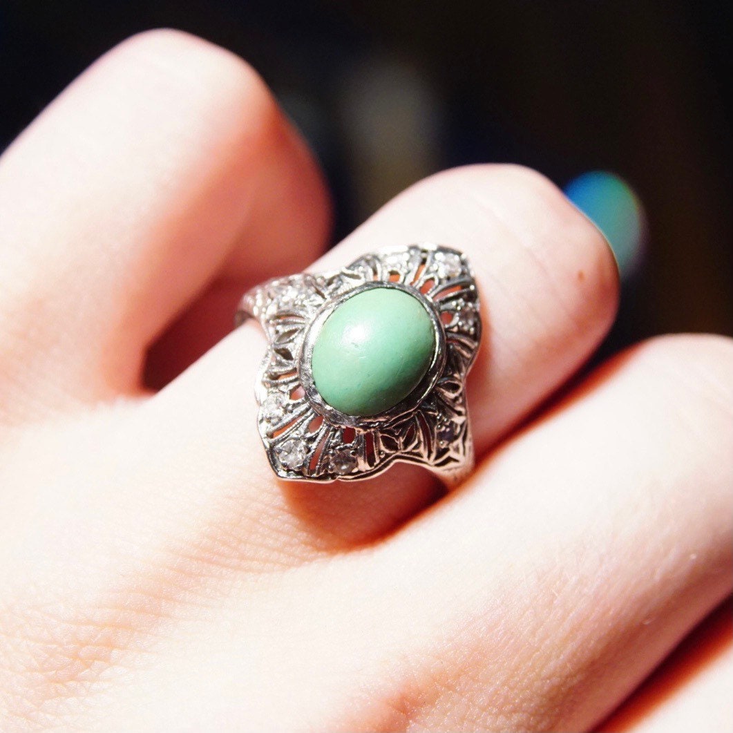 Vintage 18K white gold ring featuring a matte green turquoise stone surrounded by an intricate art deco filigree design encrusted with small diamonds, worn on a finger against a dark background.
