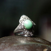 Vintage 18K white gold filigree ring with matte green turquoise center stone and diamond encrusted shield design, size 7 3/4 US, shown on dark stone surface with blurred background.